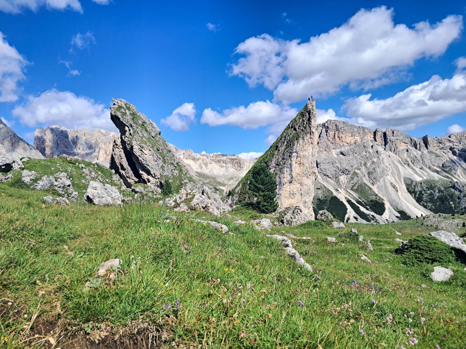 Jagged limestone rock spires at Pieralongia in Val Gardena near Seceda, surrounded by green alpine meadows and wildflowers under a bright blue sky with scattered white clouds. Two hikers stand on the pointed peak in the distance, emphasizing the dramatic scale of the Dolomite landscape.