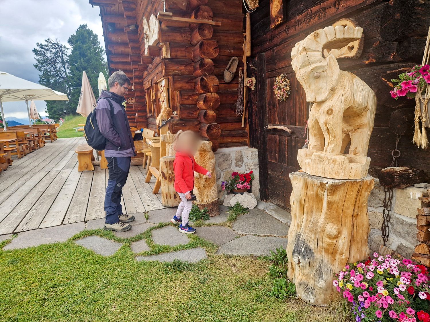 A man and child stand outside the rustic wooden Baita Seurasas mountain hut beside large hand carved wooden animal sculptures. Flower boxes and outdoor tables surround the cabin with cloudy mountain scenery in the background.