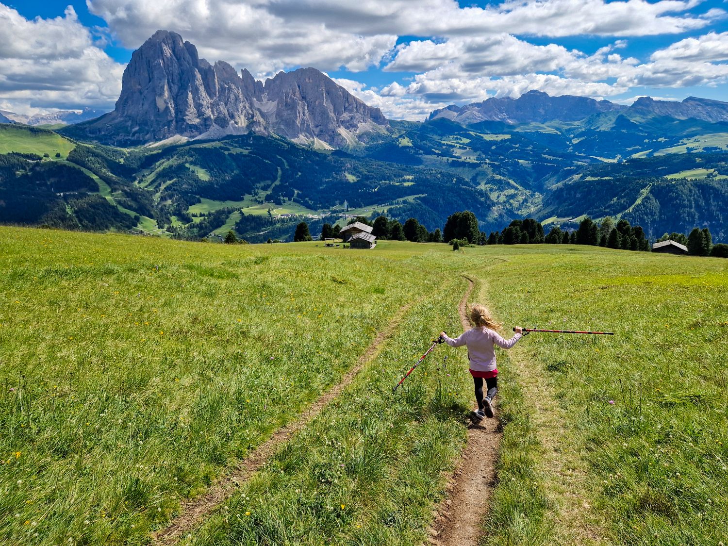 A child walks downhill with trekking poles through a wide alpine meadow while descending from Monte Pic toward the Seurasas junction. Sassolungo dominates the view across Val Gardena with scattered huts and forested slopes below.
