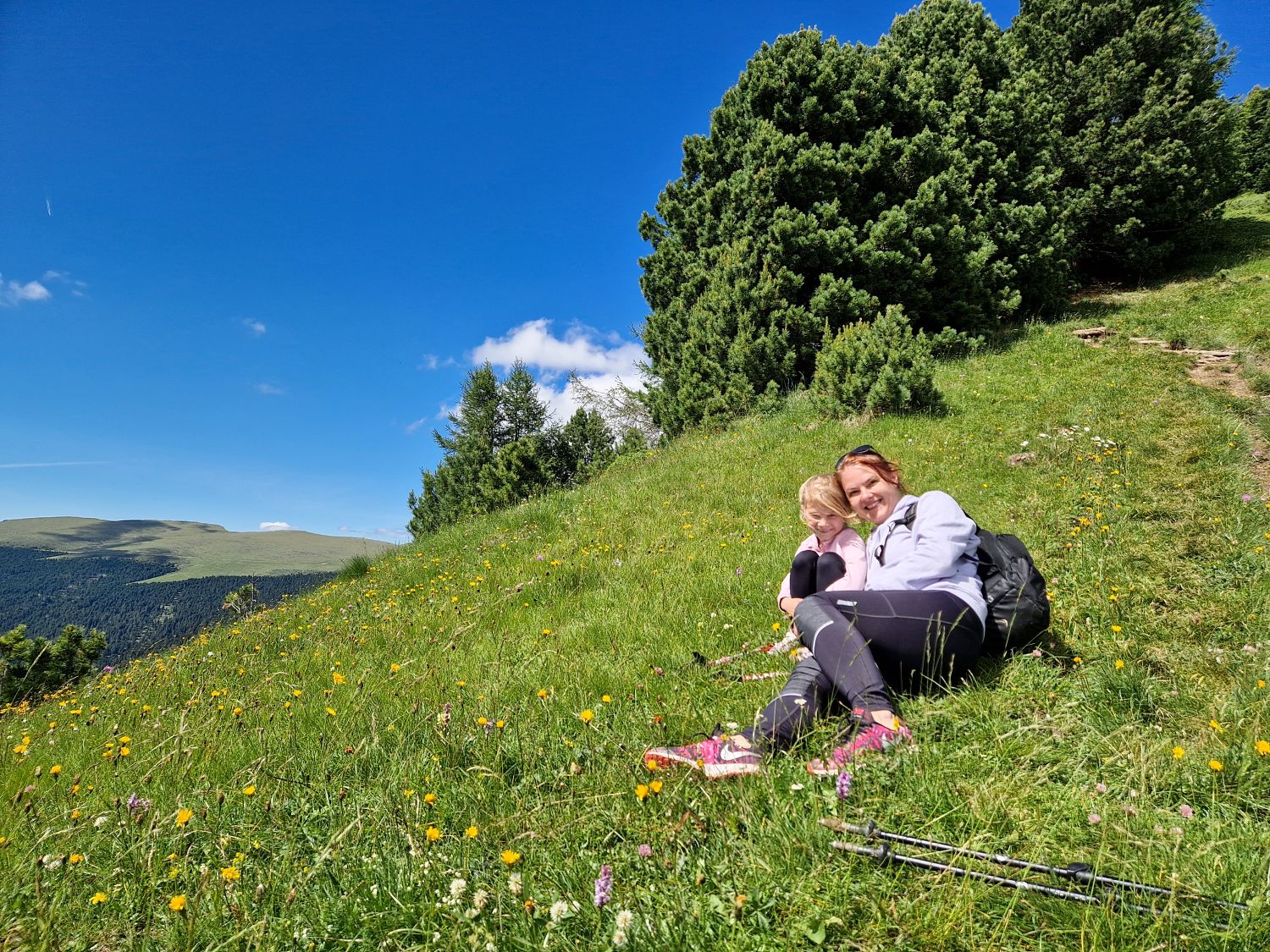 A woman and child rest in a wildflower filled meadow while descending from Monte Pic in Val Gardena. The grassy trail section sits just before the Seurasas junction with mountain pines rising behind them beneath a clear blue sky.
