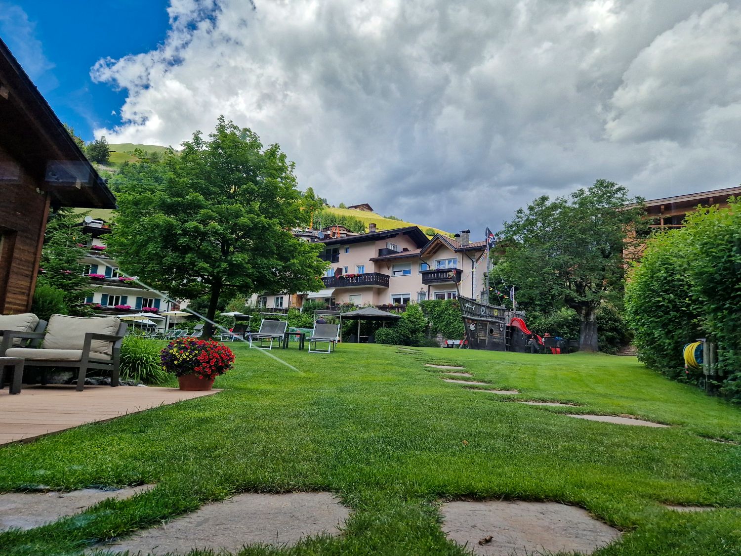 View toward Monte Pic from the garden at Hotel Posta in Santa Cristina Val Gardena. Green lawns and hotel buildings sit below the grassy mountainside beneath a dramatic cloudy sky.
