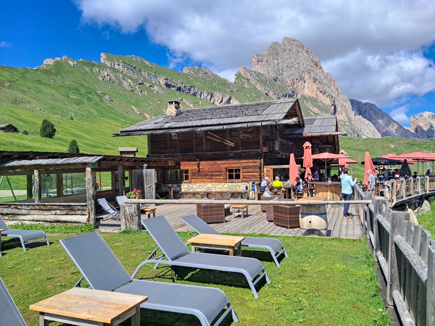 Baita Daniel mountain hut in Val Gardena with a sunny outdoor terrace, lounge chairs and hikers relaxing beneath rugged Dolomite peaks. The rustic wooden hut is a scenic stop along the Monte Pic to Col Raiser hike.
