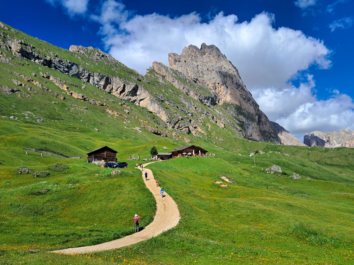 A winding gravel path leads hikers through green alpine meadows toward Baita Troier in Val Gardena. Steep rocky Dolomite slopes rise directly behind the mountain hut beneath a vivid blue sky and large white clouds.
