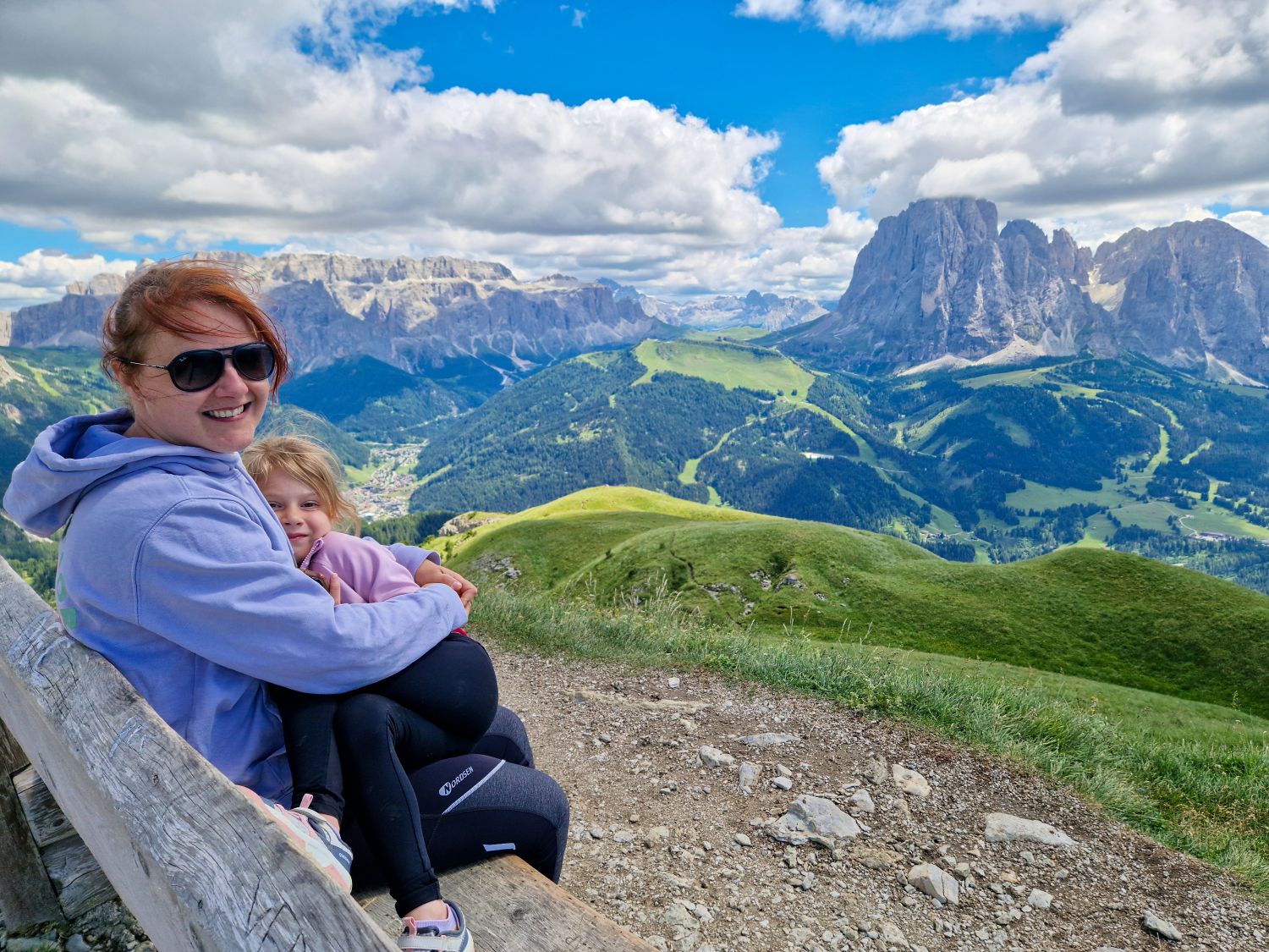 A woman and child rest on a wooden bench during the Monte Pic hike with sweeping views across Val Gardena. The green alpine ridge drops away toward the valley while the Sassolungo and surrounding Dolomite peaks rise dramatically beneath large summer clouds.
