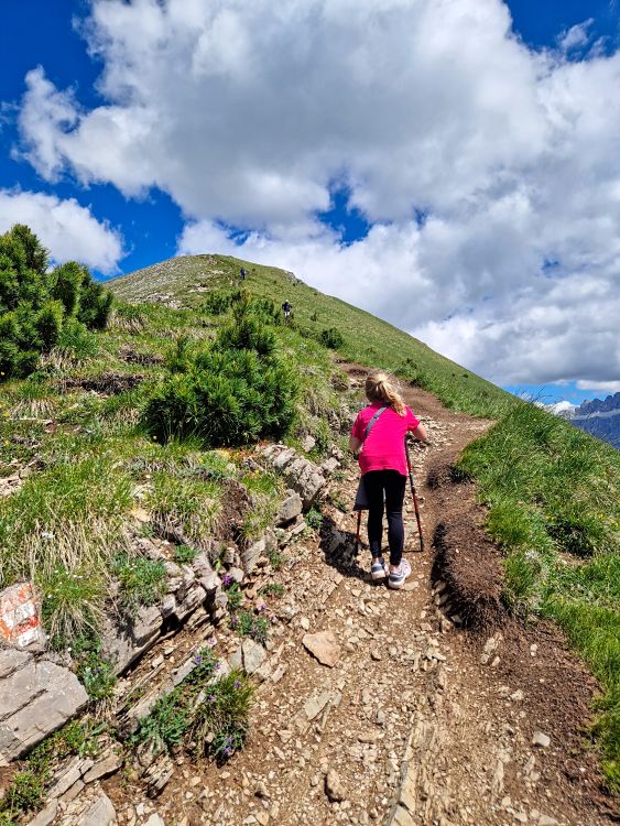 A child climbs a steep rocky section of the Monte Pic hike using trekking poles as the narrow trail winds up a grassy alpine ridge. Red and white trail markings guide the route beneath a bright blue sky filled with large clouds.
