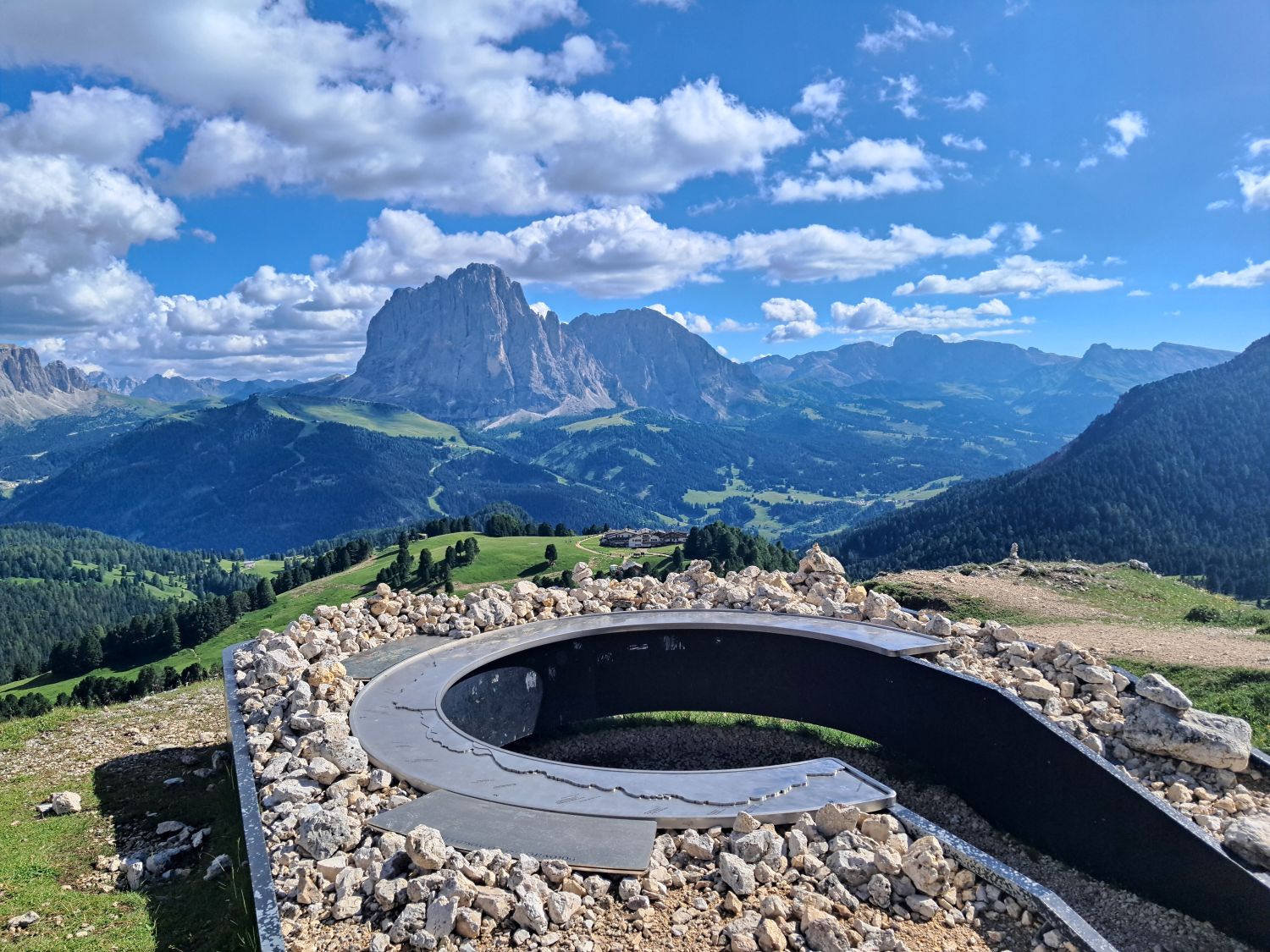 Curved viewing platform at the Mastlé UNESCO viewpoint surrounded by pale rocks and overlooking rolling alpine hills in Val Gardena. Sassolungo rises dramatically in the distance beneath a bright blue sky with scattered clouds.
