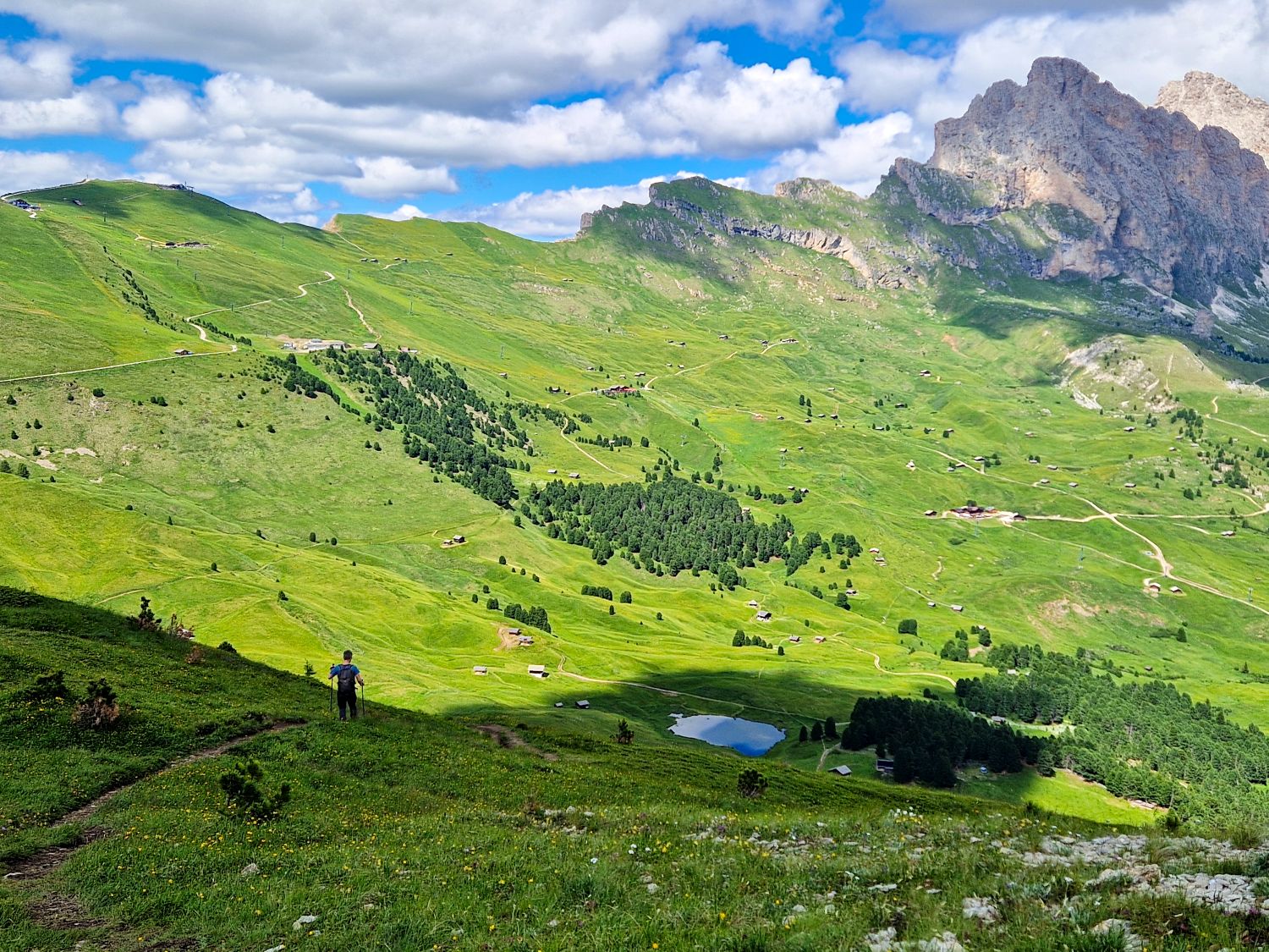 A hiker descends a narrow trail through rolling green alpine meadows on the Monte Pic hike in Val Gardena. A small mountain pond and scattered huts sit below while rugged Dolomite cliffs rise above the open landscape.

