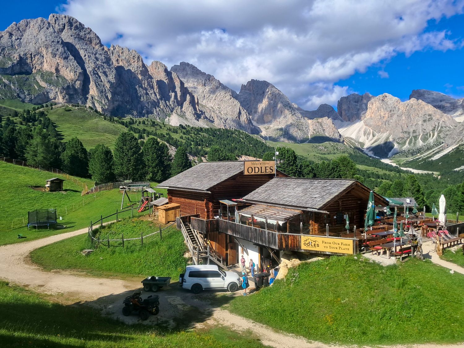 Malga Odles mountain hut sits in a green alpine meadow beneath the jagged Odle peaks in the Dolomites. The rustic wooden hut has an outdoor terrace and signs reading "Malga X Hütte ODLES" and "From Our Farm To Your Plate".
