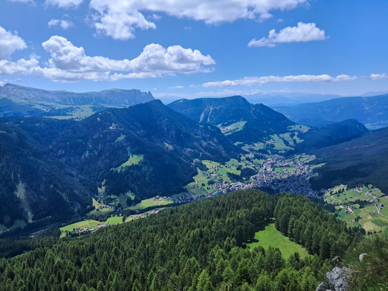Sweeping view over Val Gardena from the Monte Pic hike - Crujeta viewpoint - with forested slopes, green alpine meadows and villages spread across the valley below. Layered mountain ridges stretch into the distance beneath a bright blue sky with scattered clouds.
