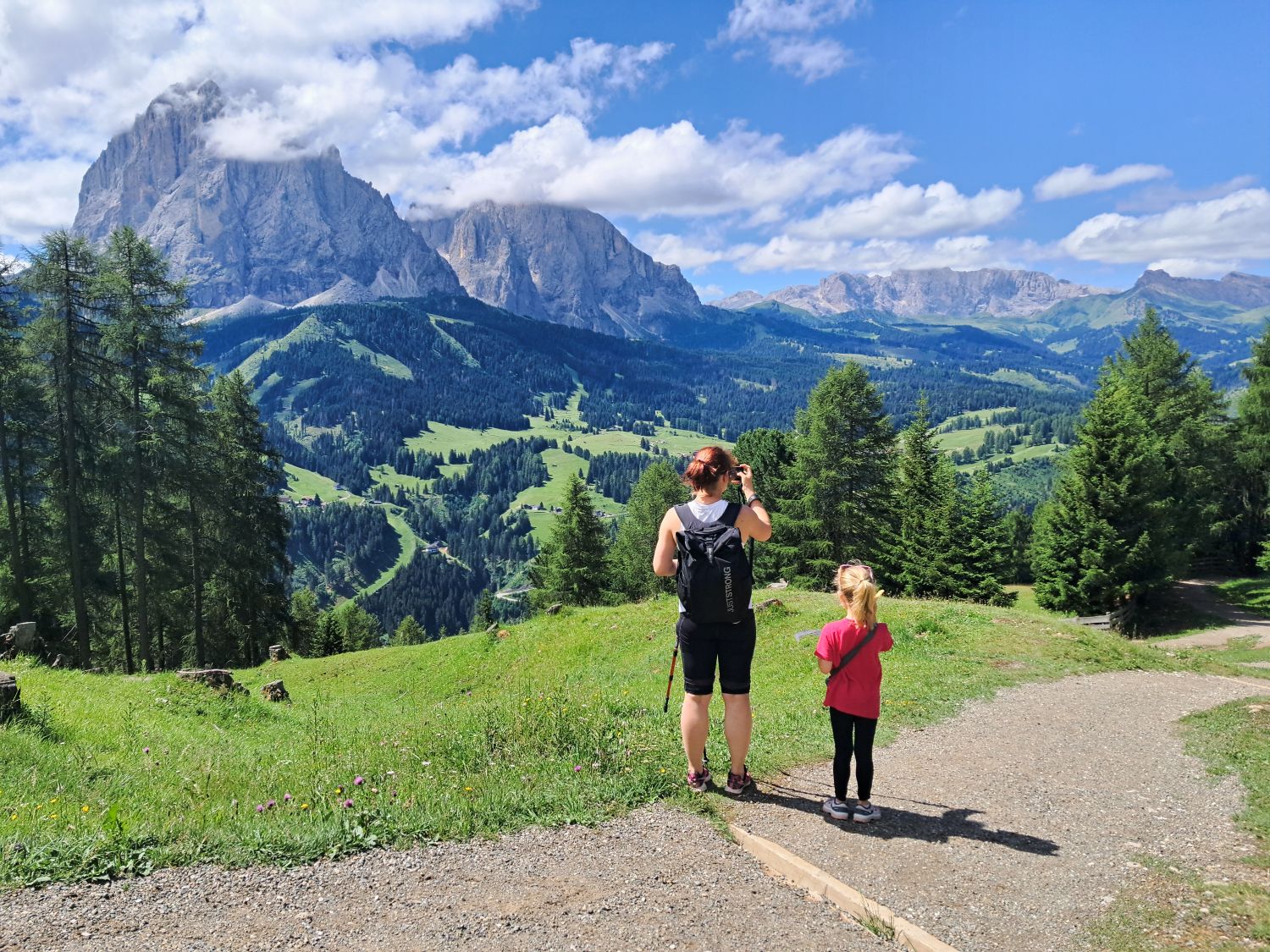 A woman and child pause on the Monte Pic hike in Val Gardena to look across green alpine meadows and forests toward dramatic Dolomite peaks. A bright blue sky and scattered clouds frame the wide mountain view near Baita Seurasas.
