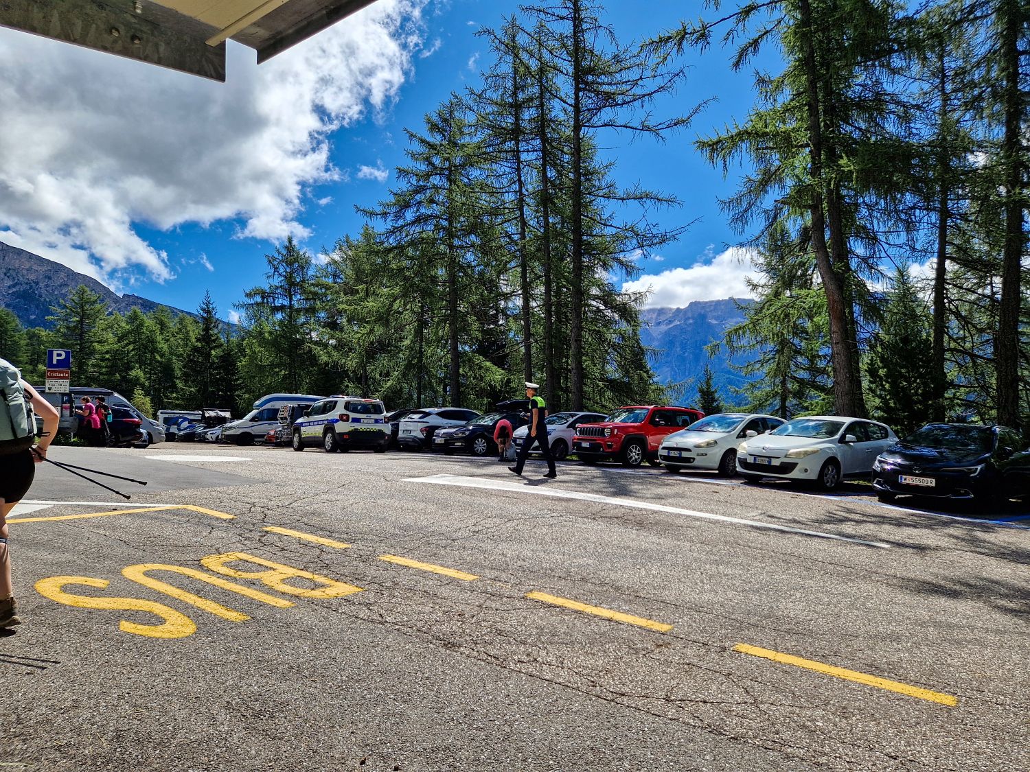Parking area at Cristauta in Val Gardena with hikers and cars surrounded by tall alpine trees. This is a common starting point for the Monte Pic hike toward Baita Seurasas with Dolomite mountains visible beyond the road.
