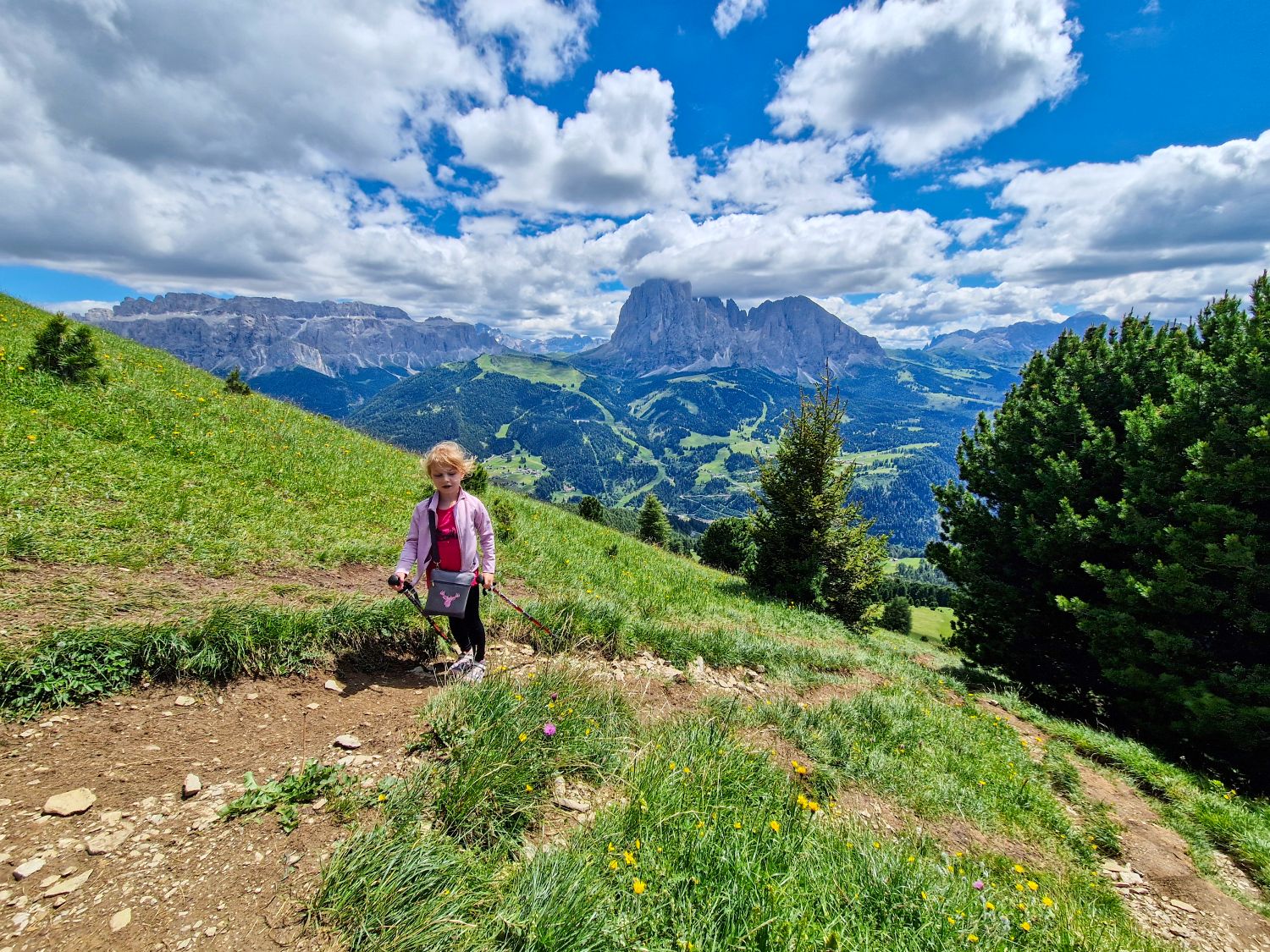 A child hikes along a narrow dirt trail through a flower filled alpine meadow on the Monte Pic hike in Val Gardena. Sassolungo and the surrounding Dolomite peaks rise dramatically across the valley beneath large summer clouds.
