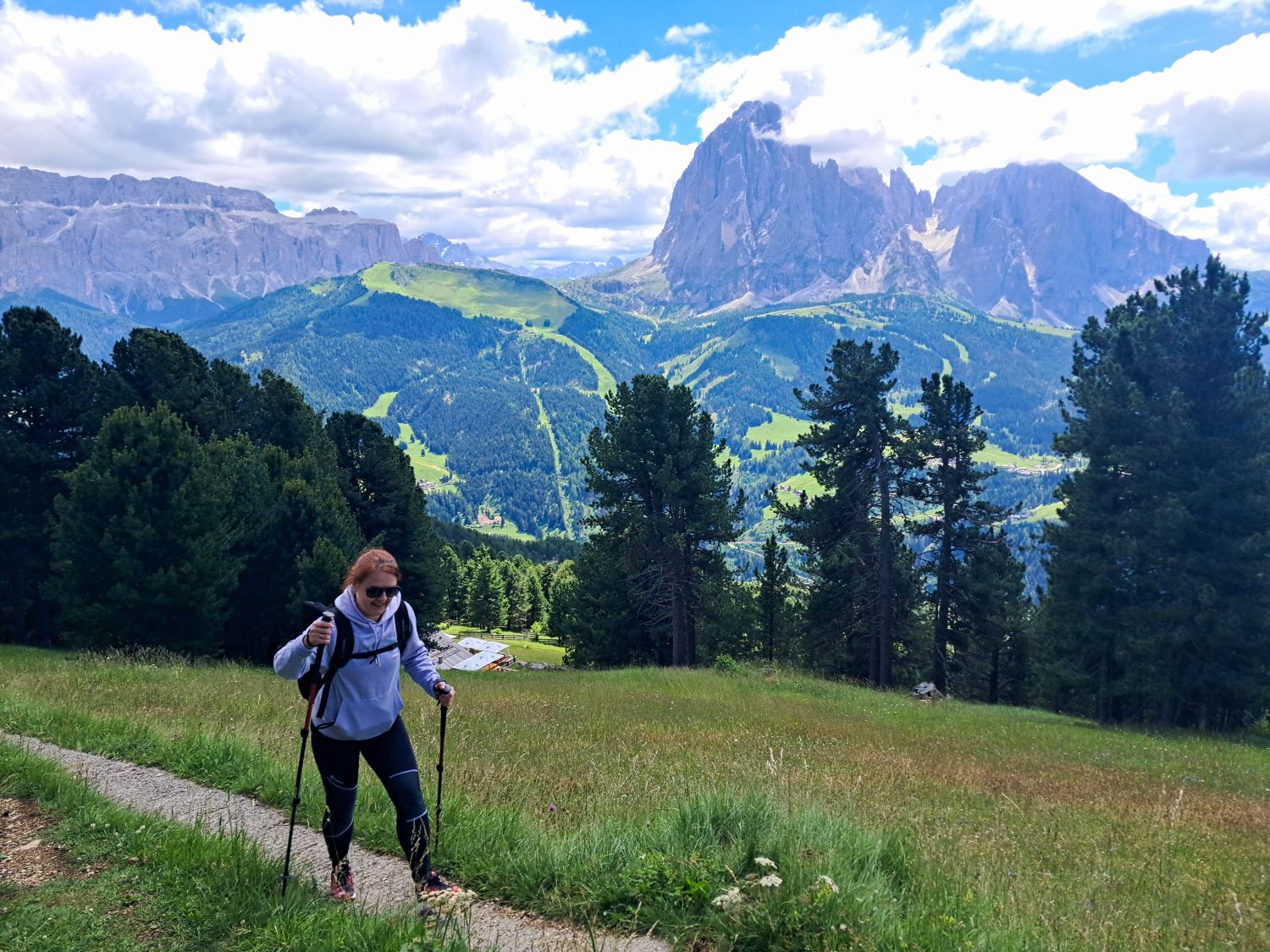 A woman hikes uphill with trekking poles through a grassy alpine meadow on the Monte Pic hike in Val Gardena. The trail opens to sweeping views of the Sassolungo and surrounding Dolomite peaks above the forested valley.
