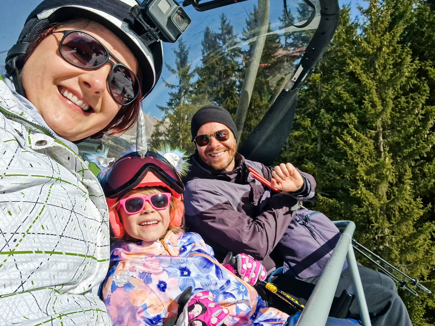 Dolomites in Winter: Weather, Skiing, Hiking, and more 2 Kristine, Emma, and Michal smile and wave while riding the Mont de Seura chairlift through snow-covered forests in the Dolomites in winter. Emma sits between Kristine and Michal wearing a helmet and pink sunglasses, while Kristine wears a helmet-mounted action camera and Michal waves from the chairlift.
