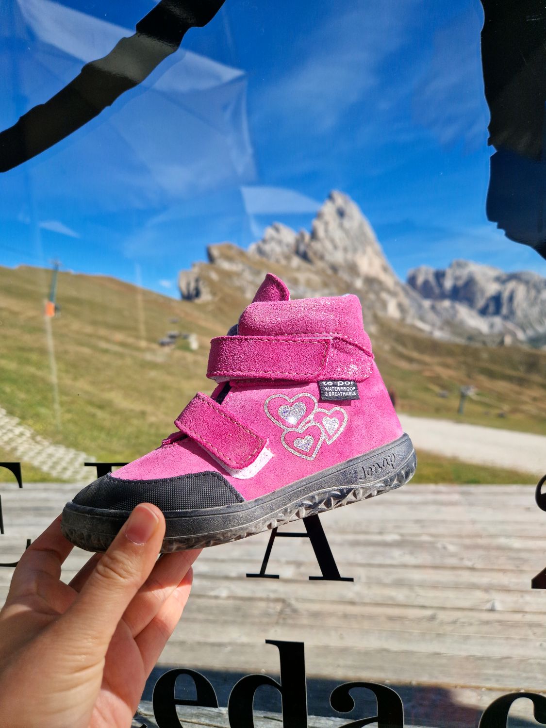 A hand holds up a bright pink kids hiking boot at Baita Sofie with the Seceda peaks and grassy slopes softly blurred in the background. The close up highlights the boot against the mountain view outside the window and the small label reads "waterproof and breathable".
