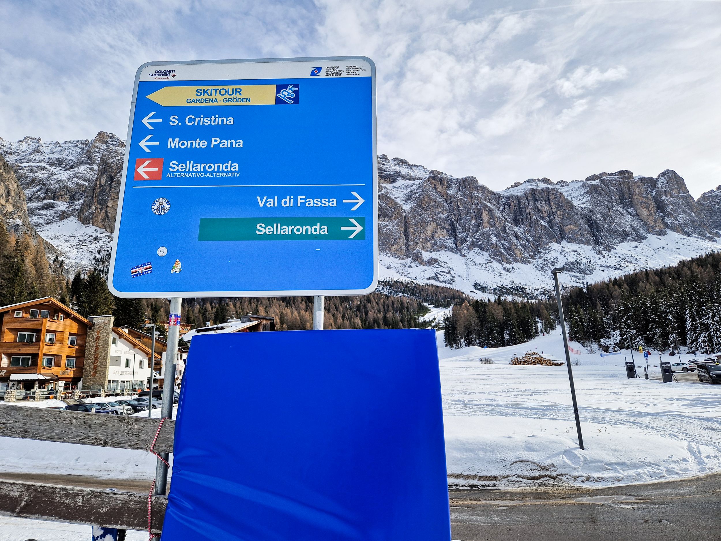 Skiing in the Dolomites for the first time 15 Blue ski route sign pointing left to S. Cristina and Monte Pana and right to Val di Fassa and the green Sellaronda route. Snow covered Dolomites mountains rise behind the sign on a winter day with ski touring and downhill skiing directions clearly marked.
